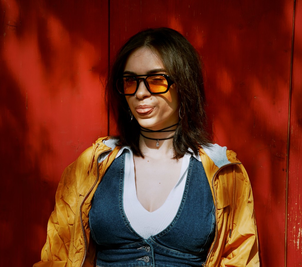 Porträt einer Frau mit orange getönter Sonnenbrille und gelber Jacke vor roter Holzwand